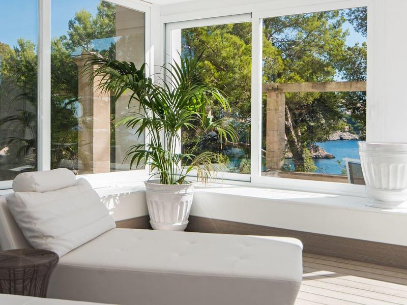 Pièce lumineuse avec grandes fenêtres, plante tropicale et vue sur la mer bleue.