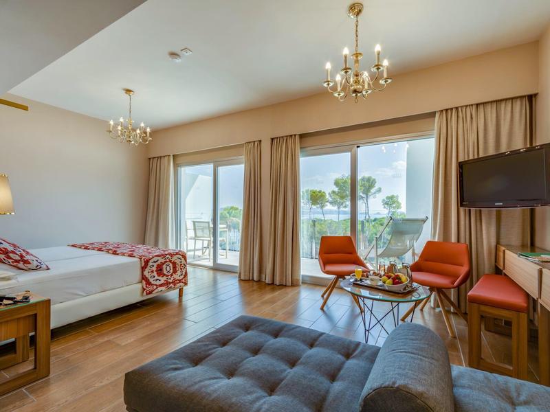 Chambre d'hôtel lumineuse avec deux lits, grandes fenêtres et coin salon avec chaises et téléviseur.