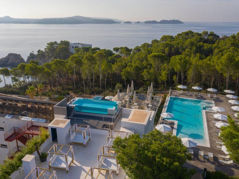 Hôtel avec grande piscine et vue sur la mer, entouré d'arbres verts et de montagnes.