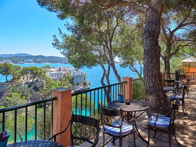 Terrasse mit Tischen und Stühlen unter Bäumen mit Blick auf das blaue Meer und Küstenhäuser.