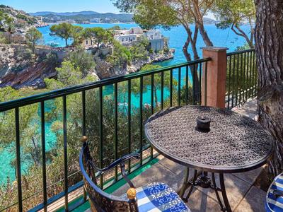 Balcón con mesa redonda y silla, vista al agua clara y costa rocosa bajo árboles.