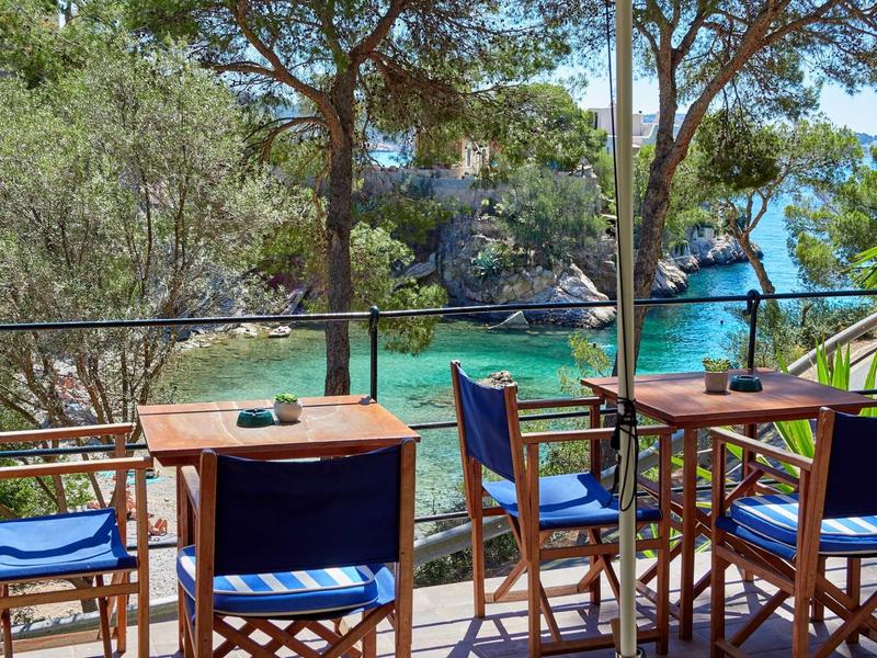 Restaurante al aire libre con mesas de madera y sillas azules frente a una costa rocosa y verde con agua clara.