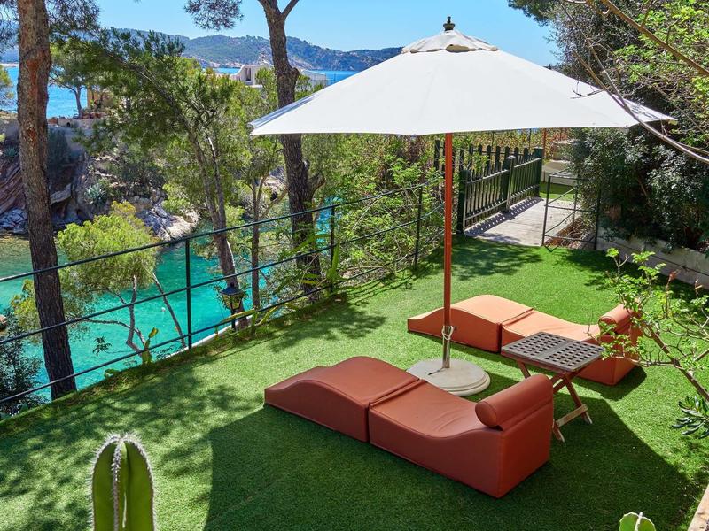 Terraza soleada con tumbonas y sombrilla, rodeada de árboles con vista al agua.