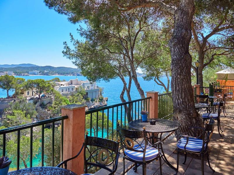 Terraza con mesas y sillas bajo árboles con vista al mar y colinas en un día soleado.