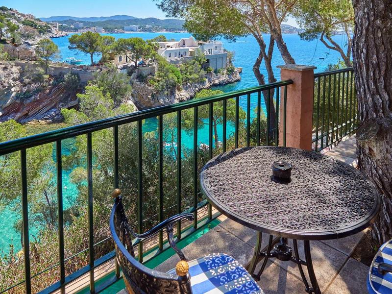 Balcón con mesa redonda y silla, vista al agua clara y costa rocosa bajo árboles.
