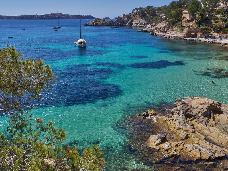 Mar turquesa claro con veleros y costa rocosa bajo colina boscosa.