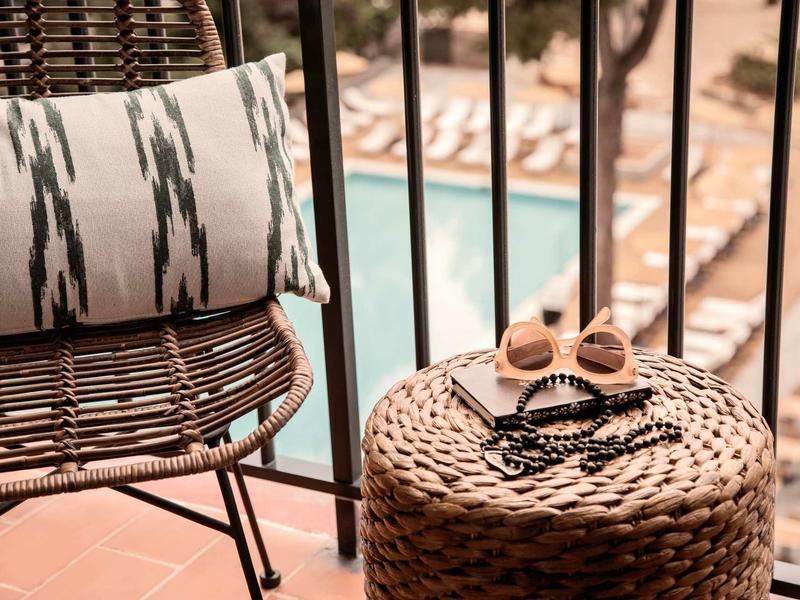 Balkon mit Stuhl und Korbtisch, darauf Sonnenbrille und Sandalen, Pool und Liegen im Hintergrund.