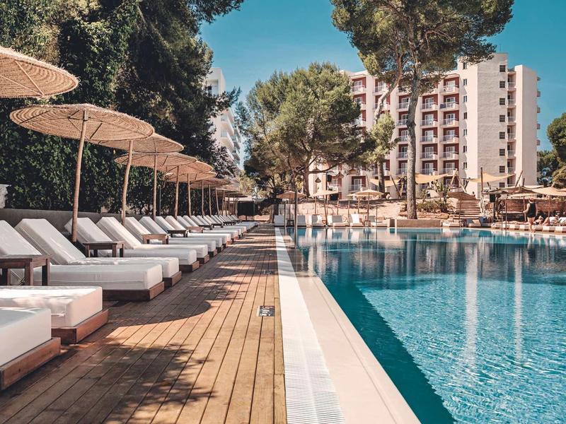 Moderner Außenpool mit Holzdeck, weißen Liegen und Sonnenschirmen vor einem mehrstöckigen Hotel.