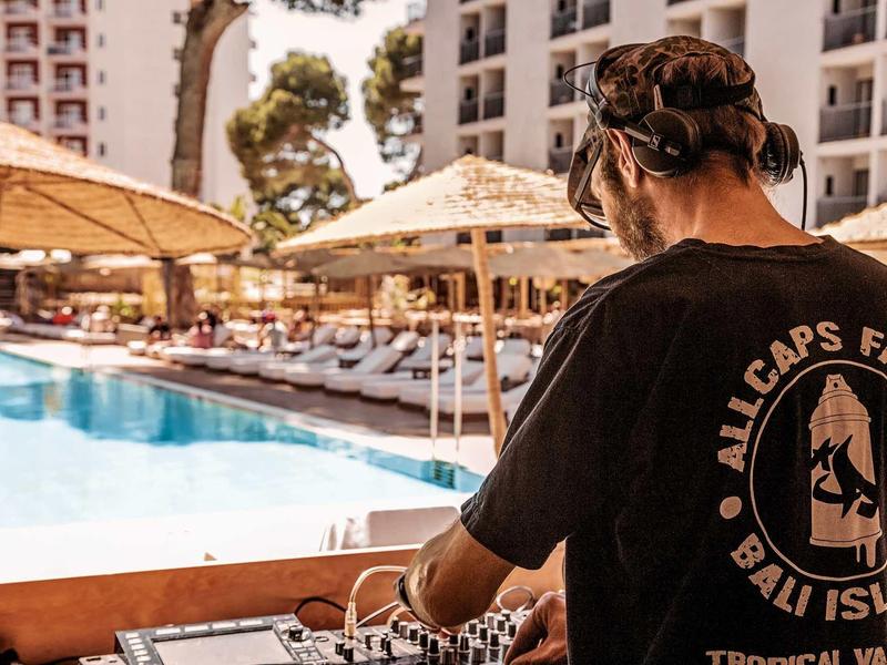 Mann mit Kopfhörern spielt Musik an Pool mit Sonnenschirmen und Hochhäusern im Hintergrund.