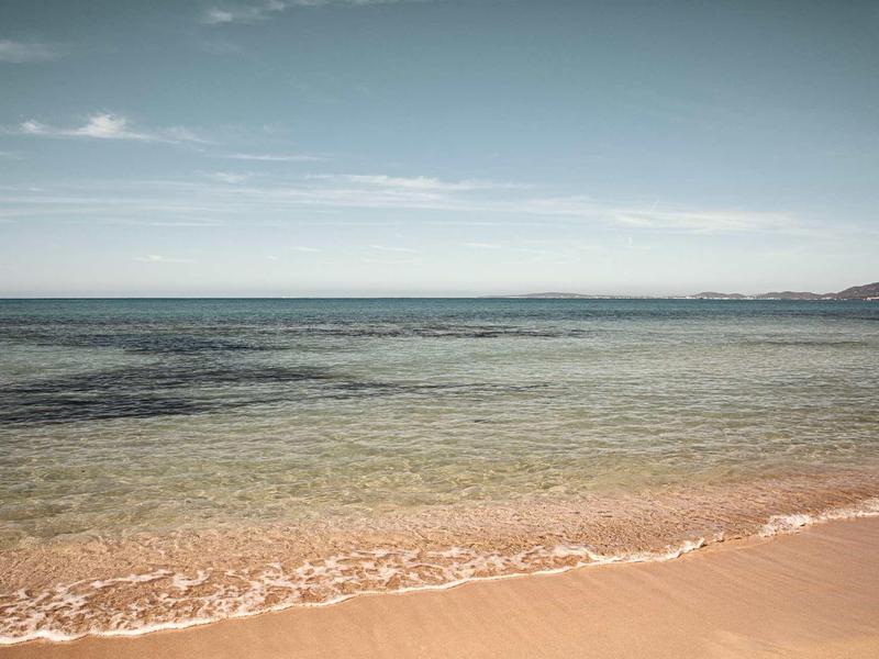 Klarer Himmel über ruhigem, flachem Meer mit sanften Wellen und hellem Sandstrand im Vordergrund.
