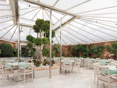 Ristorante luminoso e aperto con mobili bianchi e piante verdi sotto un tetto trasparente.