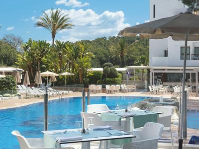Area piscina di un hotel con tavoli, sedie e ombrelloni in una giornata di sole.