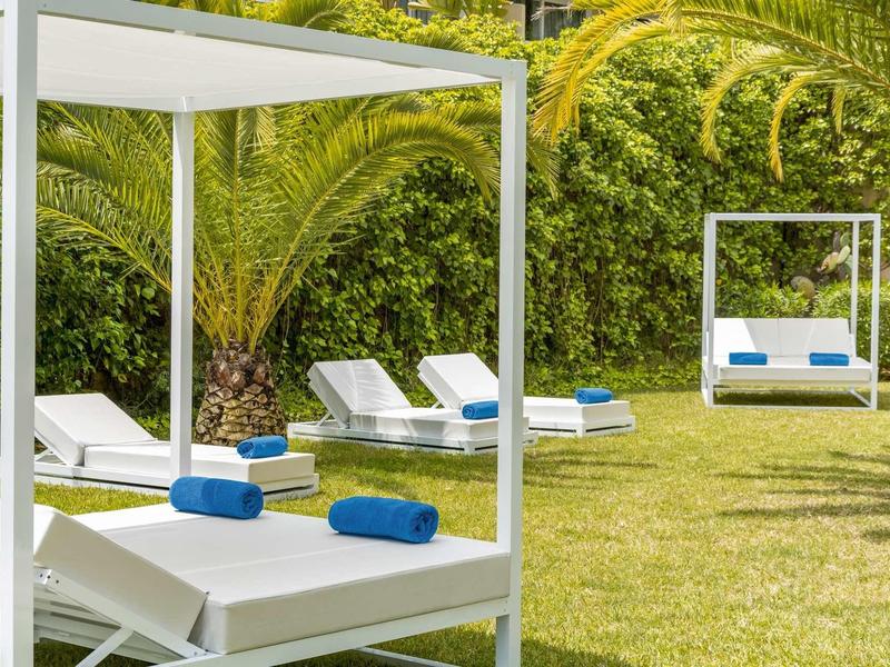 Lettini bianchi con asciugamani blu su un prato verde curato, circondati da palme.