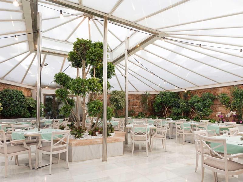 Ristorante luminoso e aperto con mobili bianchi e piante verdi sotto un tetto trasparente.