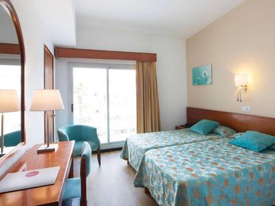 Hell eingerichtetes Hotelzimmer mit zwei Betten, Schreibtisch, Stühlen und Balkontür.