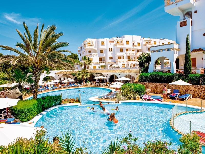 Complexe hôtelier avec piscine, palmiers et bâtiments blancs sous un ciel bleu clair