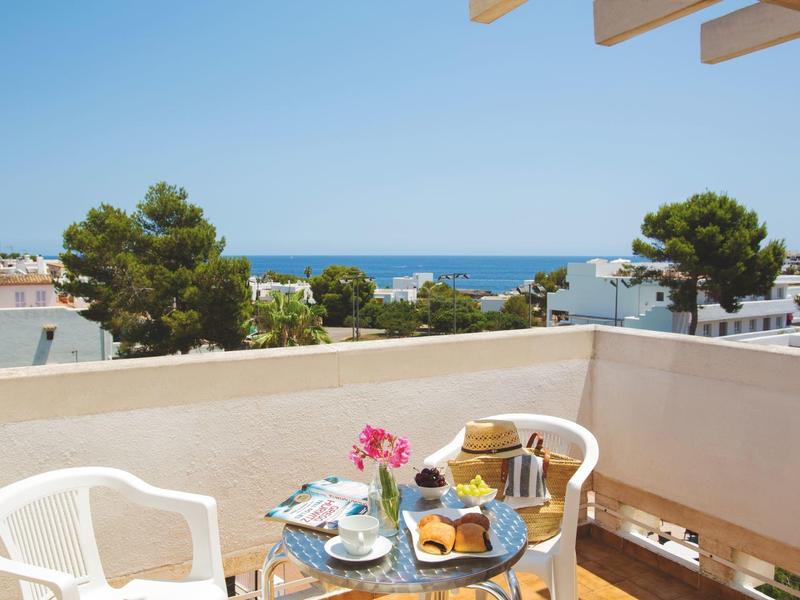 Balcon avec table de petit-déjeuner, chaises blanches et vue sur la mer et les bâtiments proches.