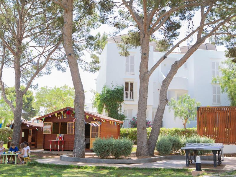 Un jardin calme avec des arbres, une cabane en bois et des bâtiments blancs en arrière-plan.