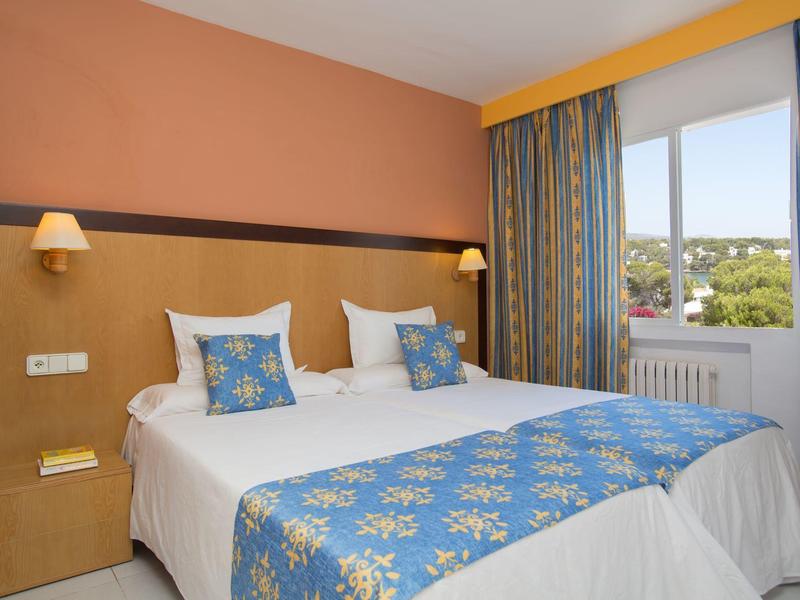 Chambre d'hôtel lumineuse avec deux lits simples, coussins colorés et grande fenêtre avec vue.