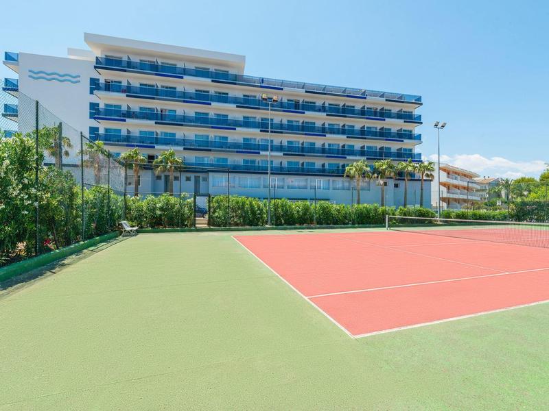 Modernes Hotel mit Tennisplatz im Freien und Palmen im Vordergrund bei klarem Himmel.