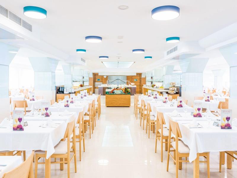 Hell beleuchteter Speisesaal mit weißen Tischen, Stühlen und blauen Deckenleuchten.