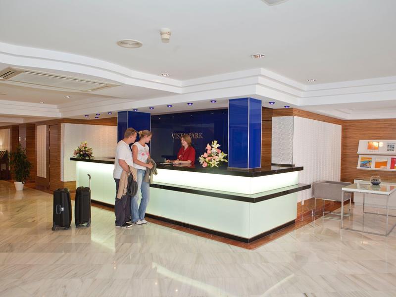 Hotelrezeption mit zwei Gästen und modernem, beleuchtetem Empfangstresen in hellem Lobbybereich.