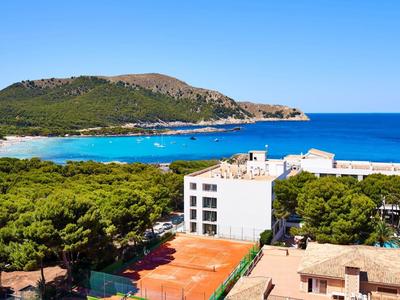 View of a coastal hotel with a tennis court, green trees, mountains, and blue sea in the background.