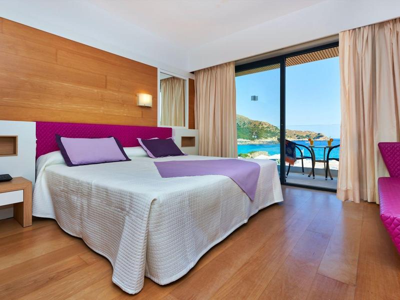 Bright hotel room with double bed, wooden floor, and balcony overlooking the sea.