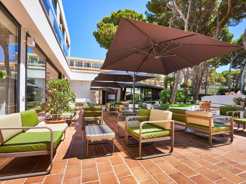 Modern hotel terrace with seating areas, sun umbrellas, and Mediterranean plants in bright sunlight.