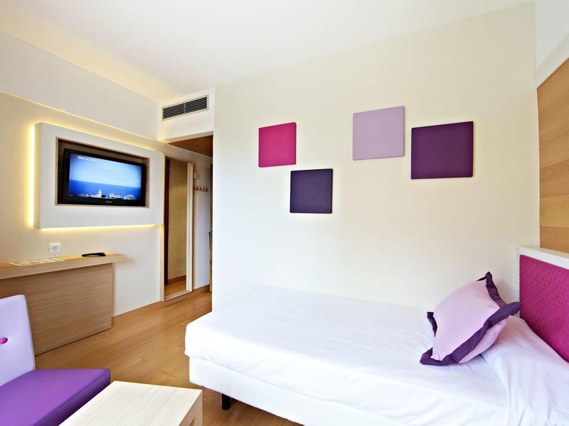 Bright hotel room with cozy bed, purple accents, and a modern wall-mounted TV.