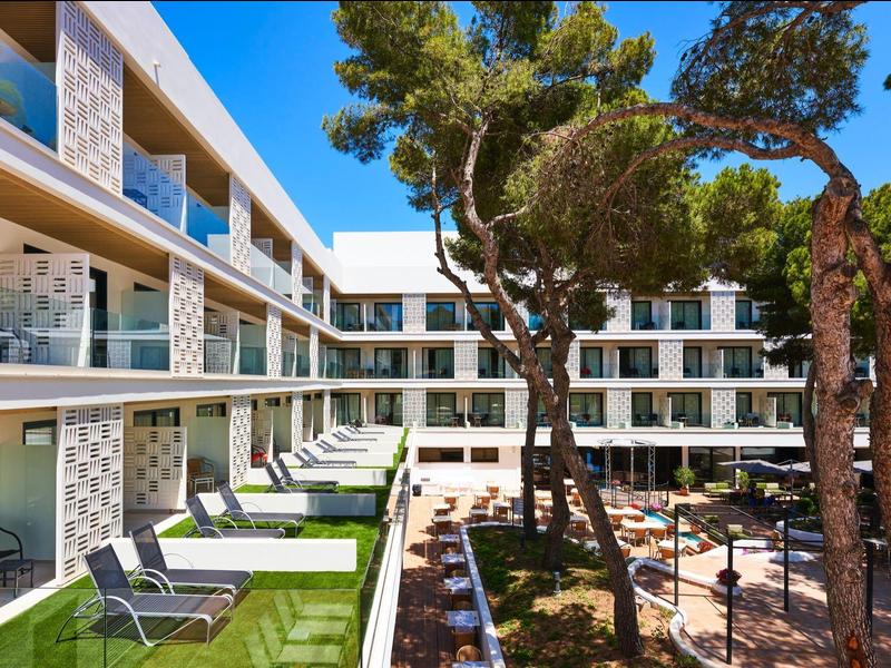 Modern hotel courtyard with lounge chairs, balconies, and large trees under a blue sky.