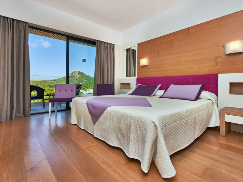 Bright hotel room with double bed, table and chair, balcony overlooking green landscape.