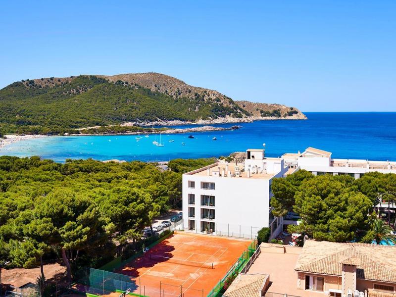 View of a coastal hotel with a tennis court, green trees, mountains, and blue sea in the background.