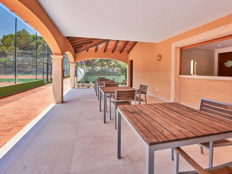 Covered terrace area with wooden tables and chairs next to a tennis court.