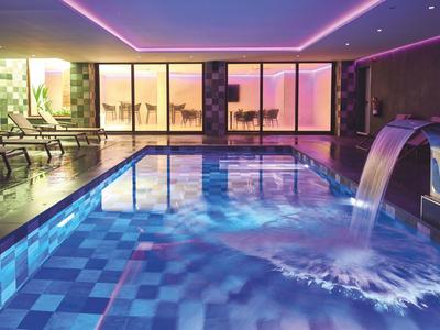 Beleuchteter Innenpool mit Wasserfall, bequemen Liegestühlen und moderner Architektur in warmem Licht.