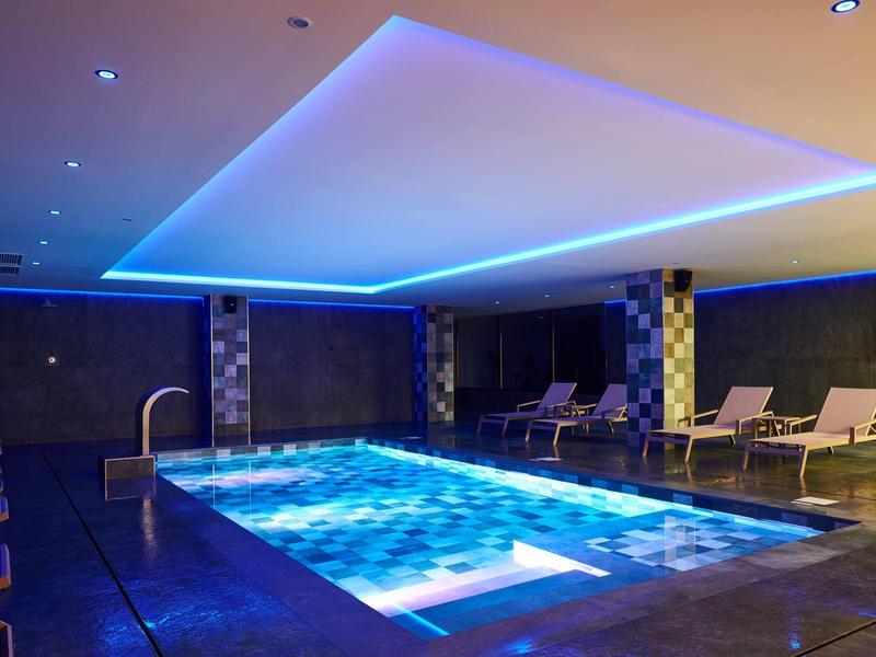 Piscine intérieure moderne avec éclairage LED bleu et chaises longues dans un cadre hôtelier élégant.
