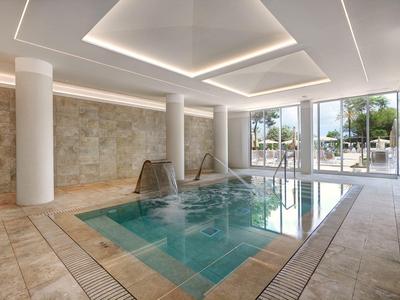 Piscina cubierta luminosa con cascada, columnas y grandes ventanales hacia el exterior.