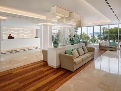 Luminosa recepción de hotel con suelo de madera y mármol, área de descanso y grandes ventanas con vistas al exterior.