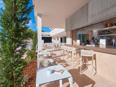 Terraza de un bar de hotel con mesas blancas, sillas y plantas verdes bajo cielo despejado