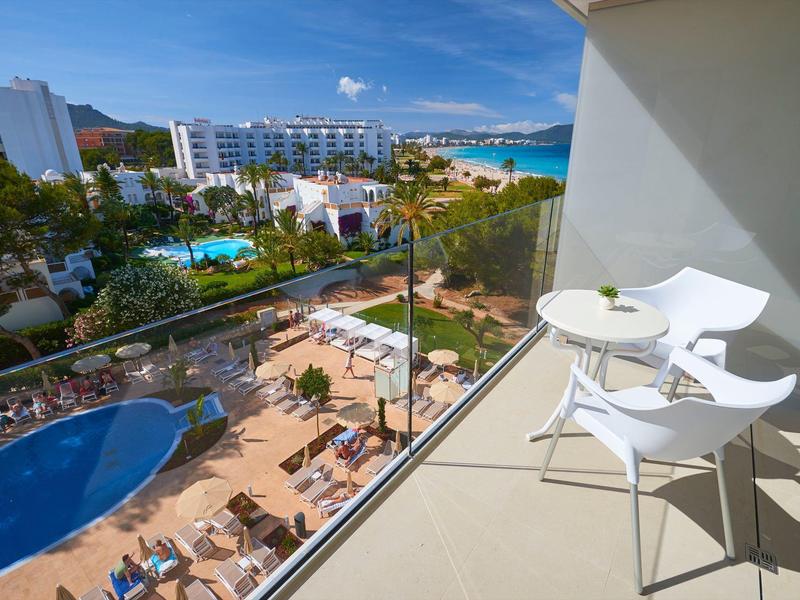Balcón con mesa y sillas blancas con vista a la piscina del hotel y al mar al fondo.