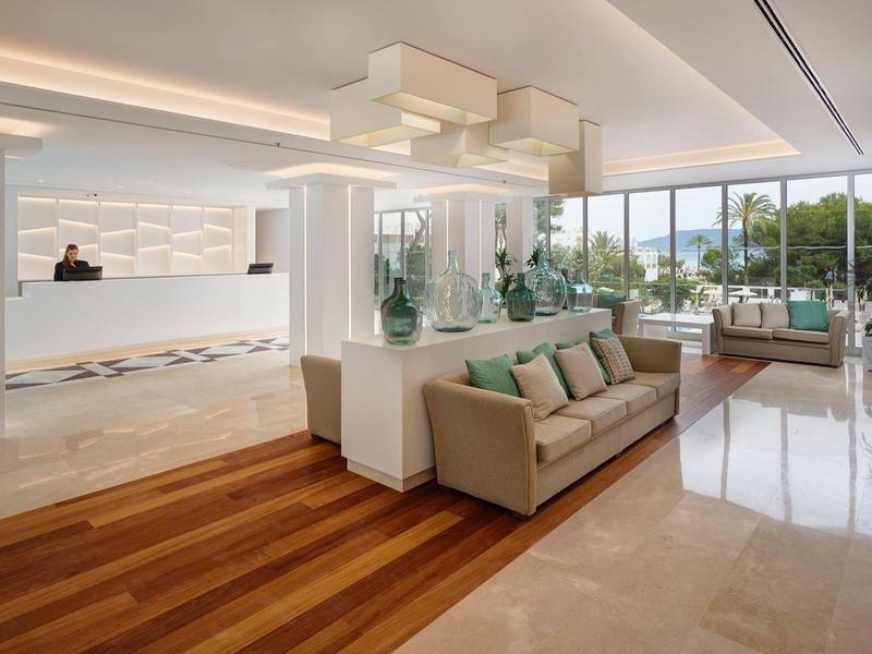 Luminosa recepción de hotel con suelo de madera y mármol, área de descanso y grandes ventanas con vistas al exterior.