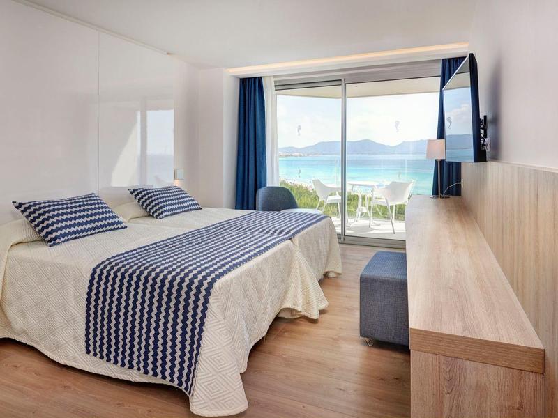 Habitación de hotel luminosa con cama doble, muebles de madera y vista al mar a través de grandes ventanas.