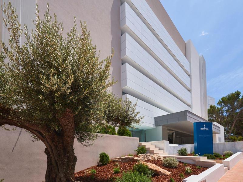 Edificio de hotel moderno de varios pisos con olivo en primer plano y cielo soleado.