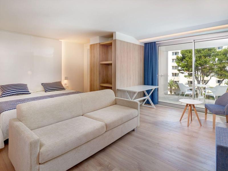 Habitación de hotel luminosa con cama doble, sofá, suelo de madera y balcón con vista a árboles.