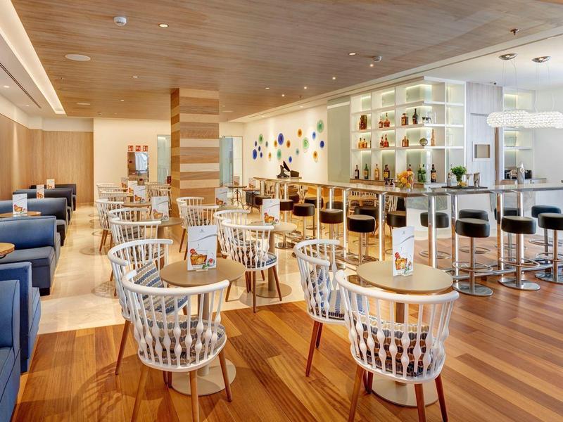 Área moderna de bar de hotel con mesas redondas, sillas blancas y taburetes en suelo de madera.