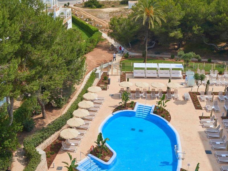 Gran piscina con palmeras y tumbonas junto a una playa de arena concurrida y mar azul.