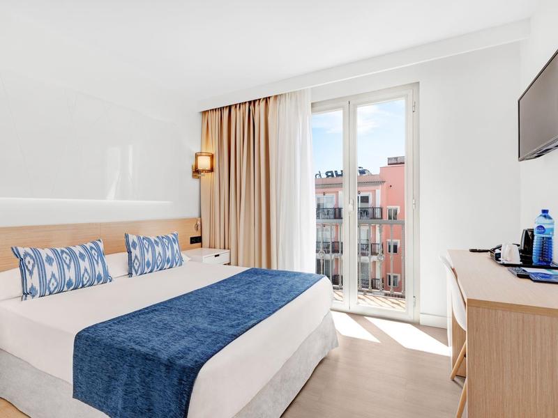 Bright hotel room with double bed, blue accents, desk, and balcony overlooking city buildings.