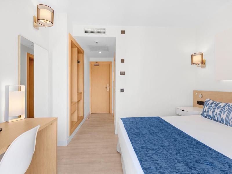 Bright hotel room with a bed, blue bedding, desk, and hallway with wooden doors.