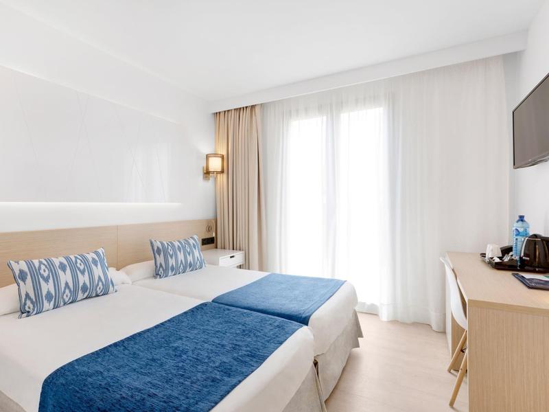 Bright hotel room with twin beds, blue accents, wall TV, desk, and balcony doors with curtains.