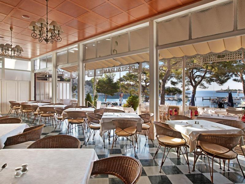 Indoor dining area with tables, wicker chairs, and large windows overlooking a tree-lined waterfront.
